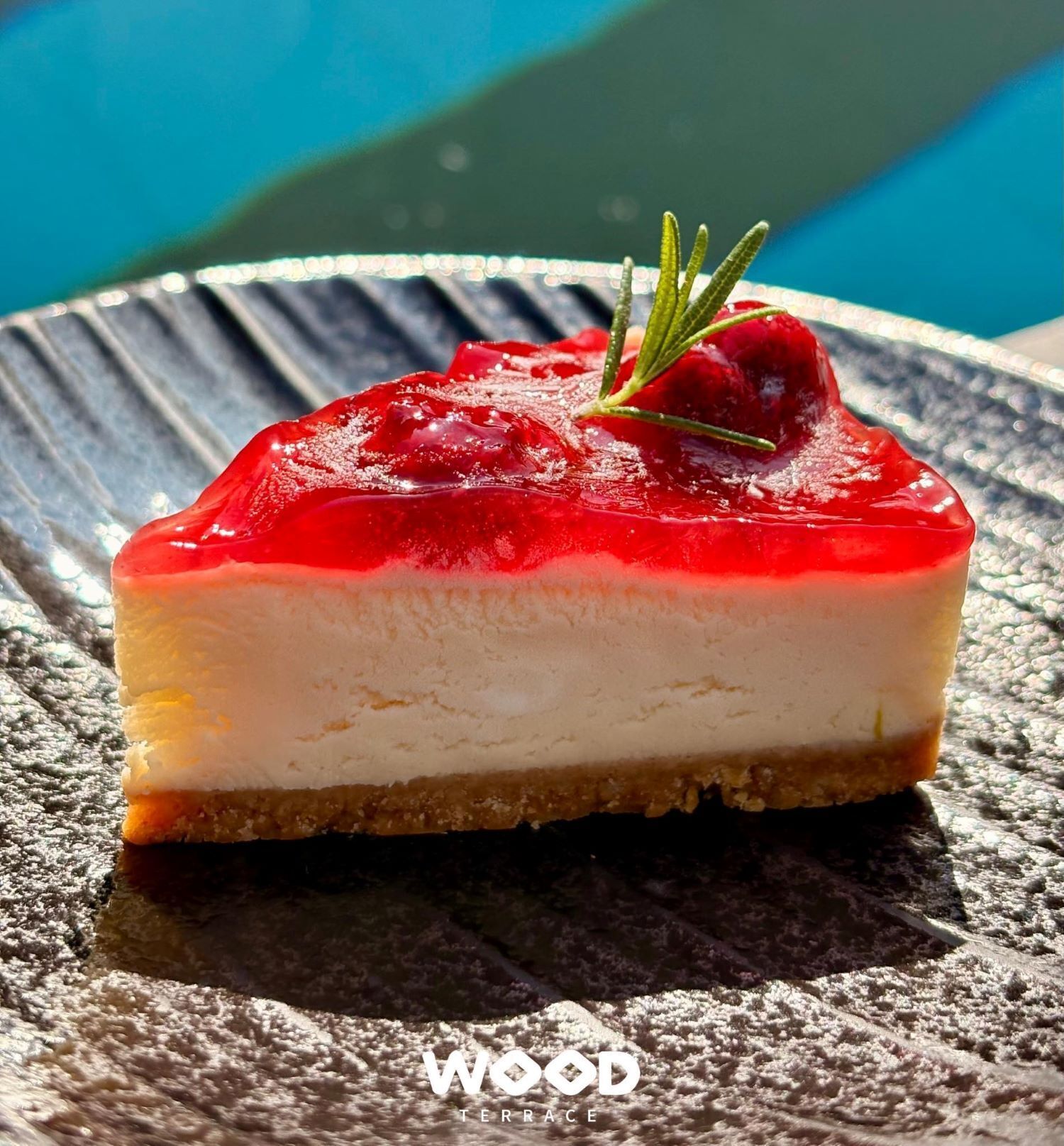 Ein Stück Käsekuchen mit roter Glasur und einem Rosmarinzweig auf einer strukturierten Platte.