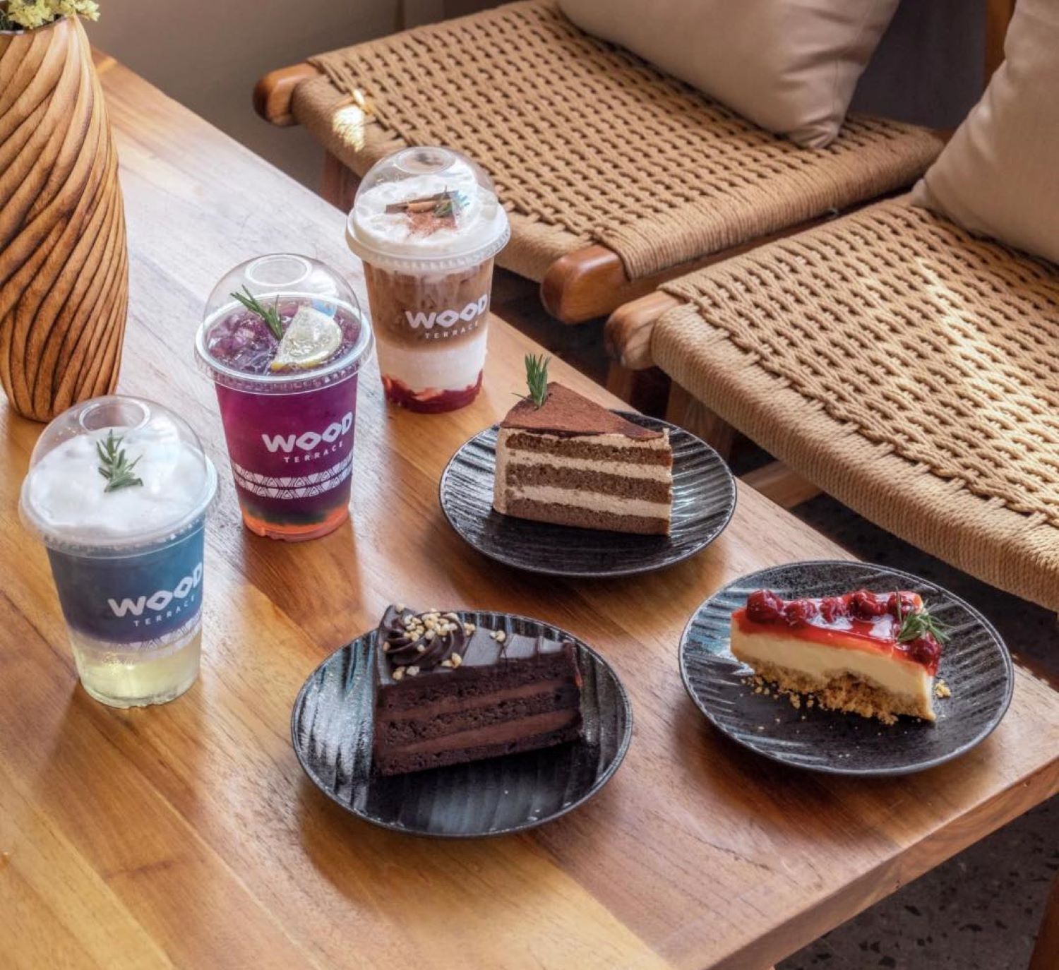 Una mesa de madera con diversos postres y bebidas, incluyendo un pastel en capas, un postre de chocolate y dos bebidas con la etiqueta 'Wood Terrace'.