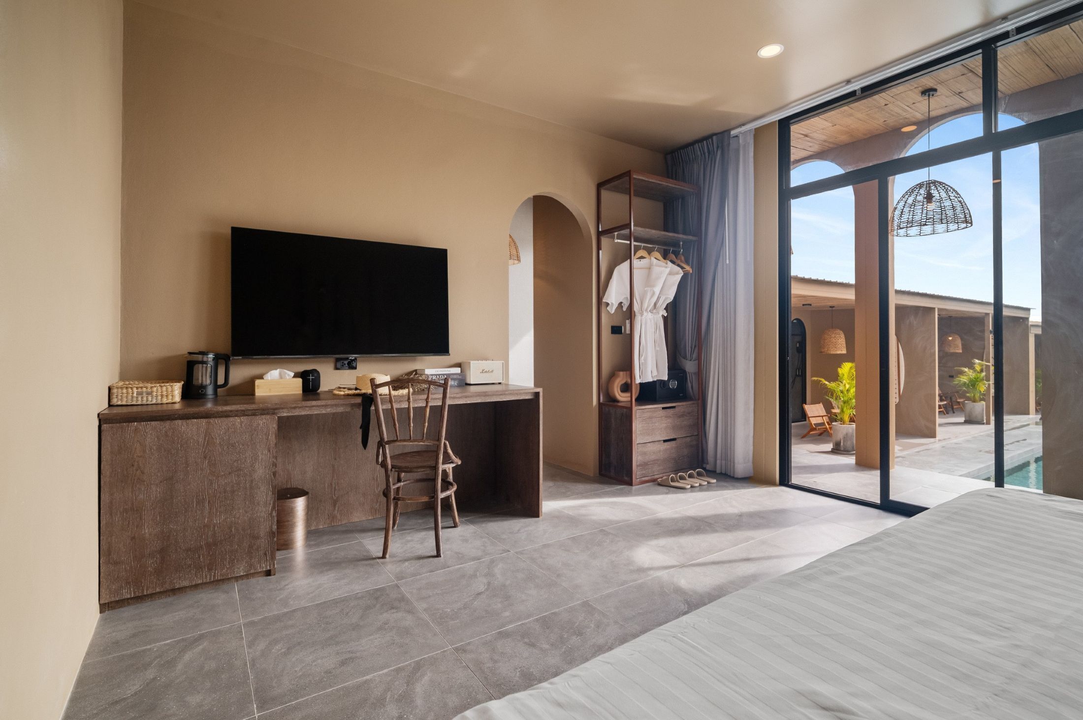 Une chambre d’hôtel moderne équipée d’un téléviseur à écran plat, d’un bureau en bois, d’une chaise, d’une armoire et d’une grande porte-fenêtre coulissante donnant sur un espace extérieur agrémenté de plantes et d’une piscine.