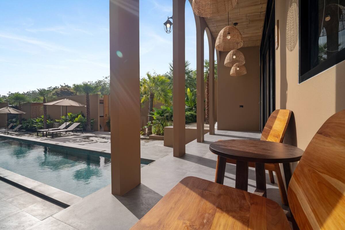 Un espace aménagé au bord de la piscine, avec une table et des chaises en bois, un luminaire suspendu et une vue sur la piscine et la végétation environnante.