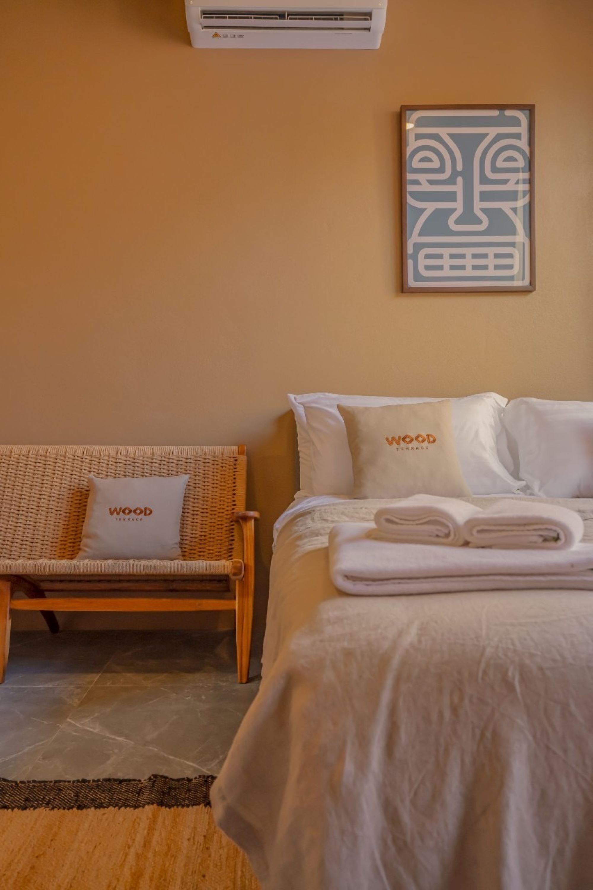 A hotel room with a bed, a wicker chair, and a wall-mounted air conditioner.