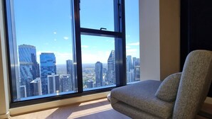 A room with a large window offering a city view, a grey armchair, and wooden flooring. - Melbourne CBD | High Rise Traveller's Paradise (Melbourne)