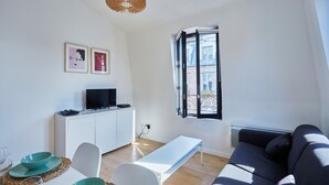 Una sala de estar moderna con un sofá, una mesa de centro, un comedor con mesa y sillas, un televisor sobre un mueble bajo y una ventana con vista a edificios.