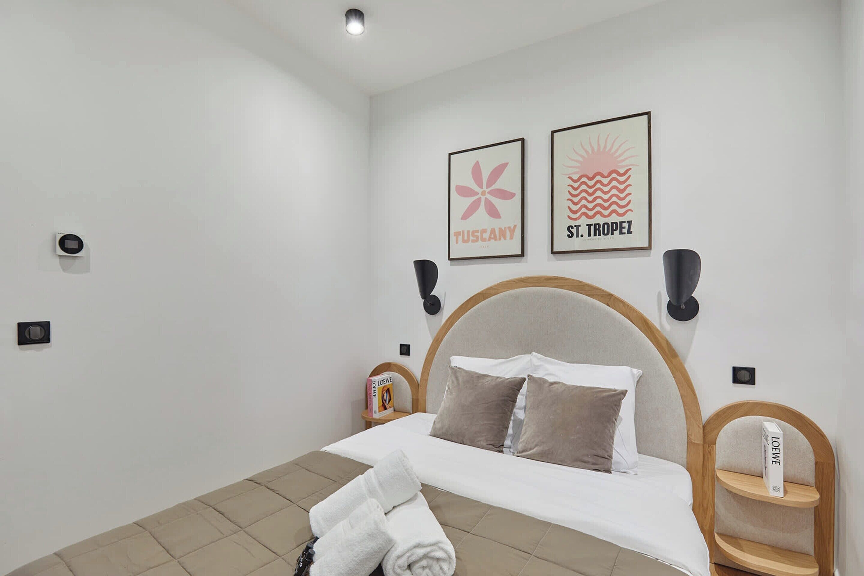 A neatly made bed with white linens, a wooden headboard, and two pillows. There are two wall-mounted speakers, a framed picture of Tuscany, and another of St. Tropez. A small bedside table with a book and a decorative item is also visible.