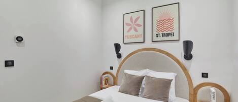 A neatly made bed with white linens, a wooden headboard, and two pillows. There are two wall-mounted speakers, a framed picture of Tuscany, and another of St. Tropez. A small bedside table with a book and a decorative item is also visible.