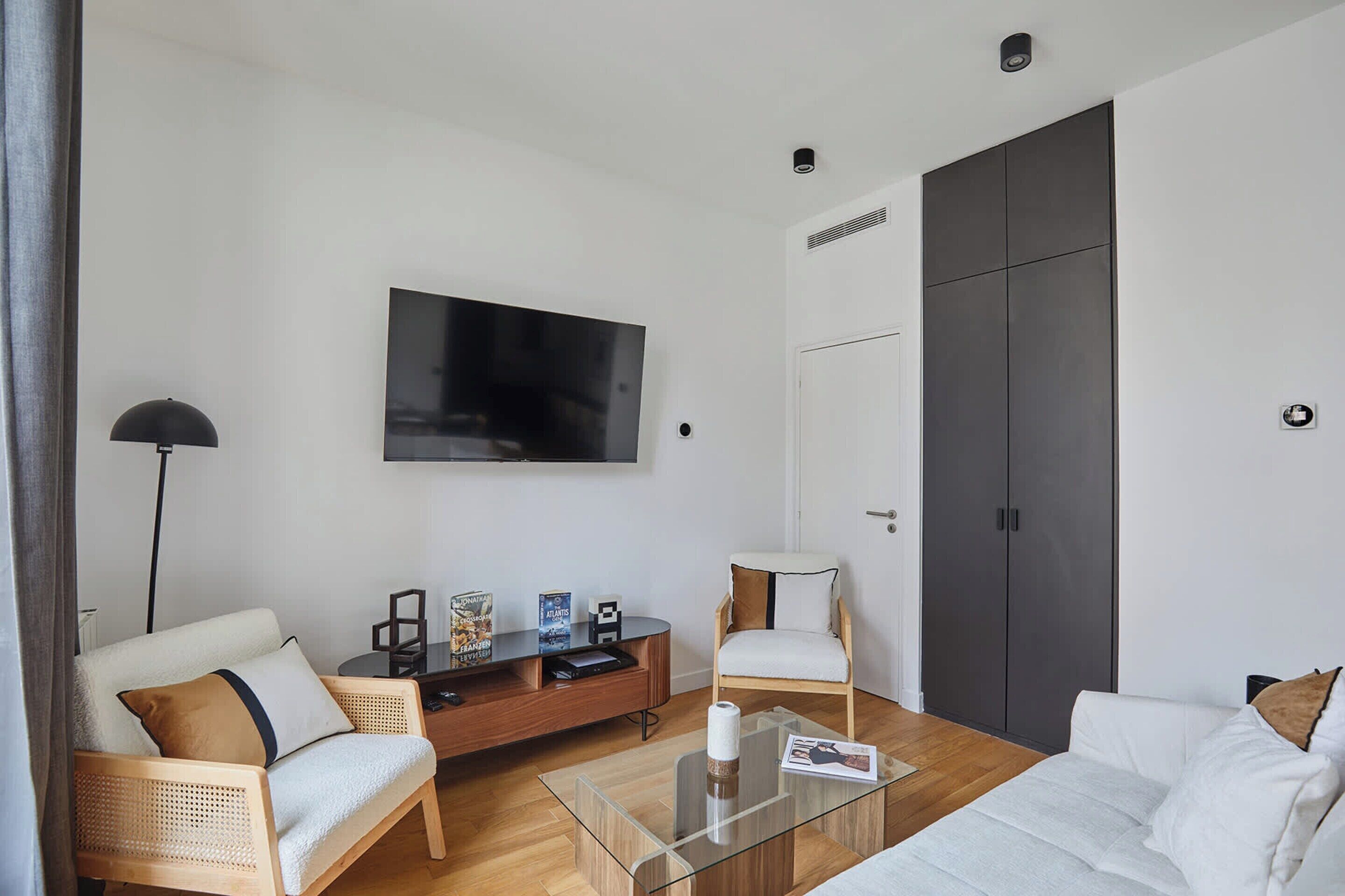 Een moderne woonkamer met een bank, fauteuil en salontafel, een flatscreen televisie aan de muur bevestigd, en een donkere deur.