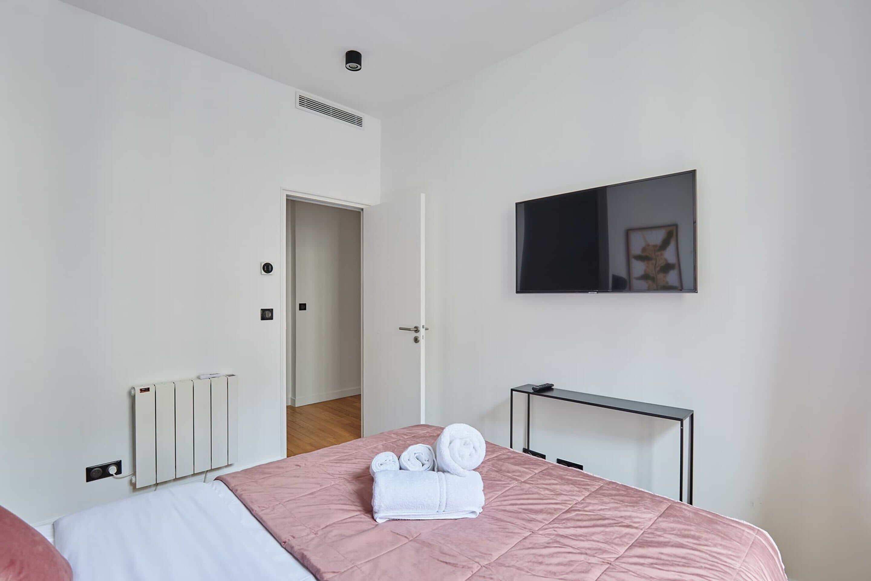 Een moderne hotelkamer met een bed, een aan de muur bevestigde televisie, een radiator en een deur die naar een andere kamer leidt.