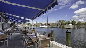 Un espace de restauration en plein air, avec des tables et des chaises sous une marquise bleue, offrant une vue sur le bord de l’eau, avec des bateaux et des bâtiments en arrière-plan.