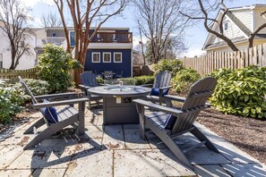 Eine Terrasse mit einem runden Tisch und vier Adirondack-Stühlen, umgeben von einem Holzzaun und verschiedenen Pflanzen.
