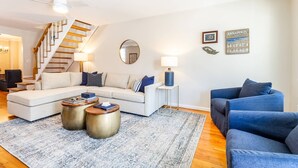 Un salon moderne comprenant un canapé, deux tables d’appoint, une table basse, un fauteuil bleu, un escalier et un mur orné d’un miroir et de deux panneaux.
