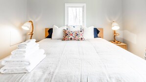 Un lit bien fait, avec des draps blancs, un coussin coloré et une pile de serviettes blanches posée sur le sol.