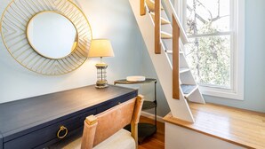 Une chambre moderne avec un bureau en bois foncé, un miroir rond, une lampe, une chaise, une petite table ronde et un escalier en bois menant à un balcon d’où l’on peut admirer la vue sur les arbres.