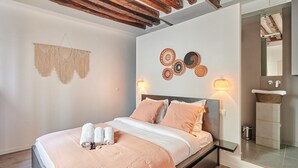 A modern bedroom with a wooden ceiling, a bed with pink bedding, wall art, and a bathroom visible through an open door. - Superb Parisian Apartment Center of Paris (Paris)