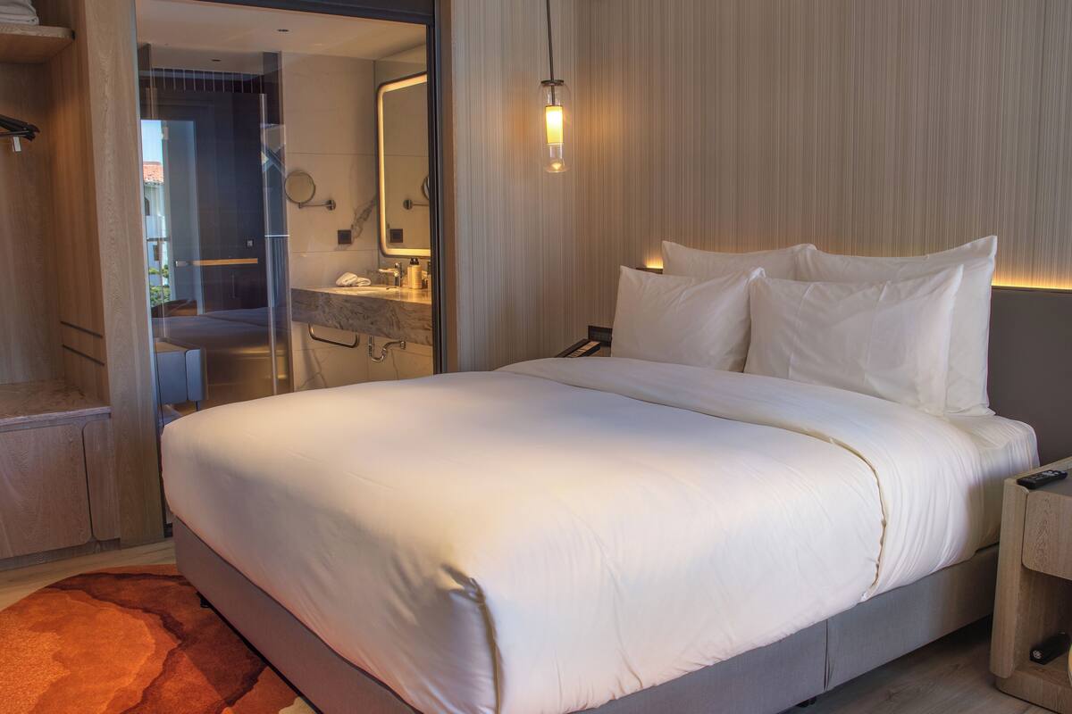 A modern hotel room with a large bed, a bathroom with a mirror, and a wooden nightstand.