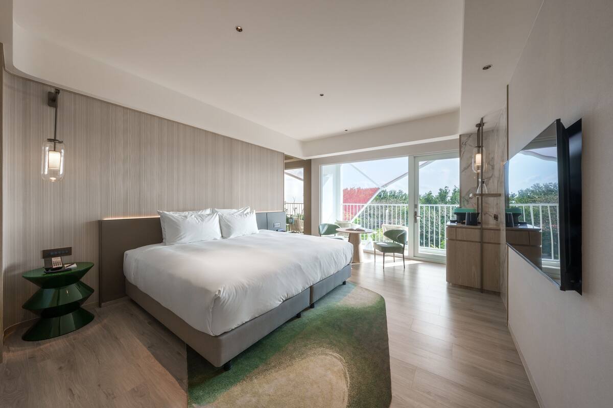 A modern bedroom with a large bed, a green side table, a balcony with a view, and a mounted TV.