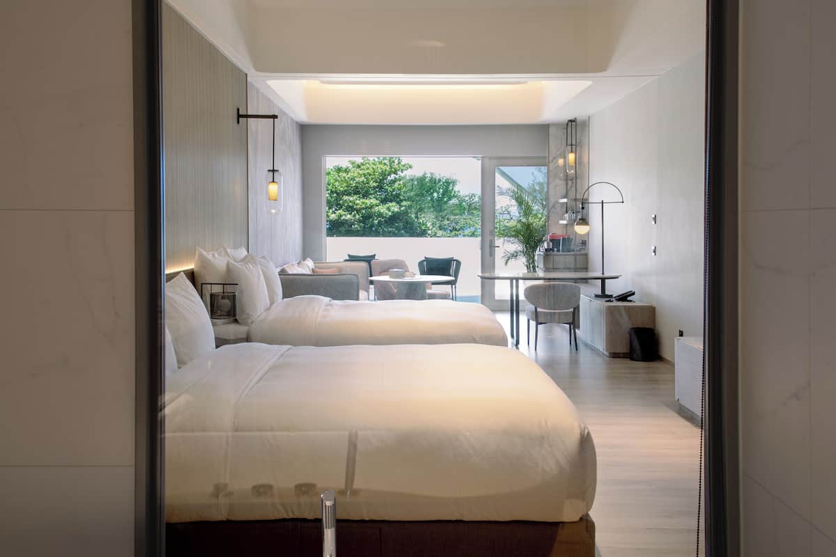 A modern hotel room with a large bed, a sitting area with a table and chairs, a small kitchen area, and a view of the outdoors.