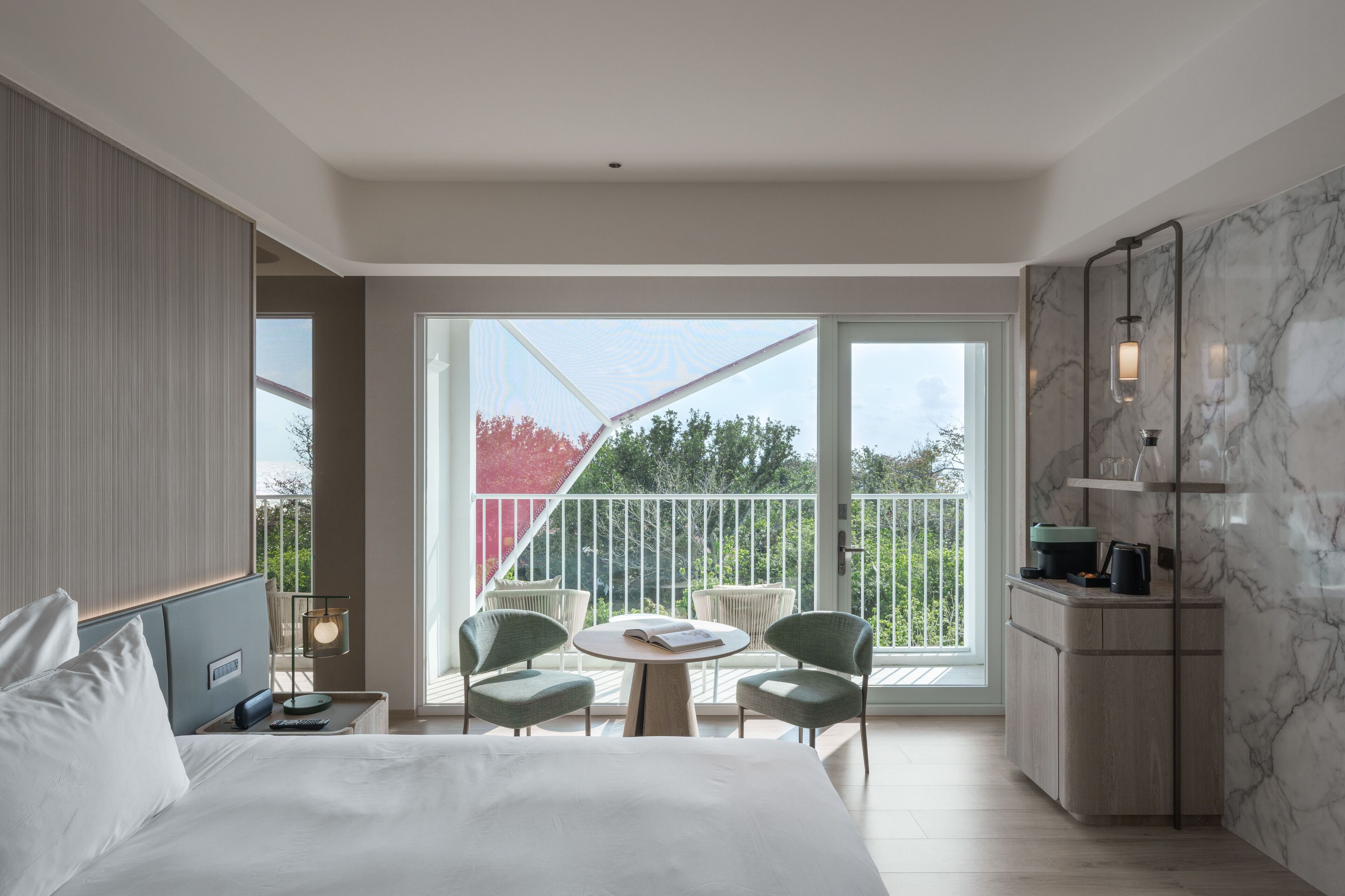 Een moderne hotelkamer met een groot bed, een kleine tafel met stoelen, een balkon met uitzicht op bomen en een kitchenette.
