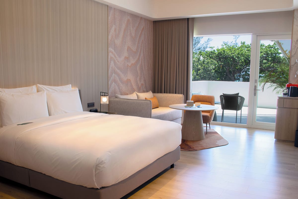 A modern hotel room with a large bed, a sofa, a small table, and a view of the outdoors.