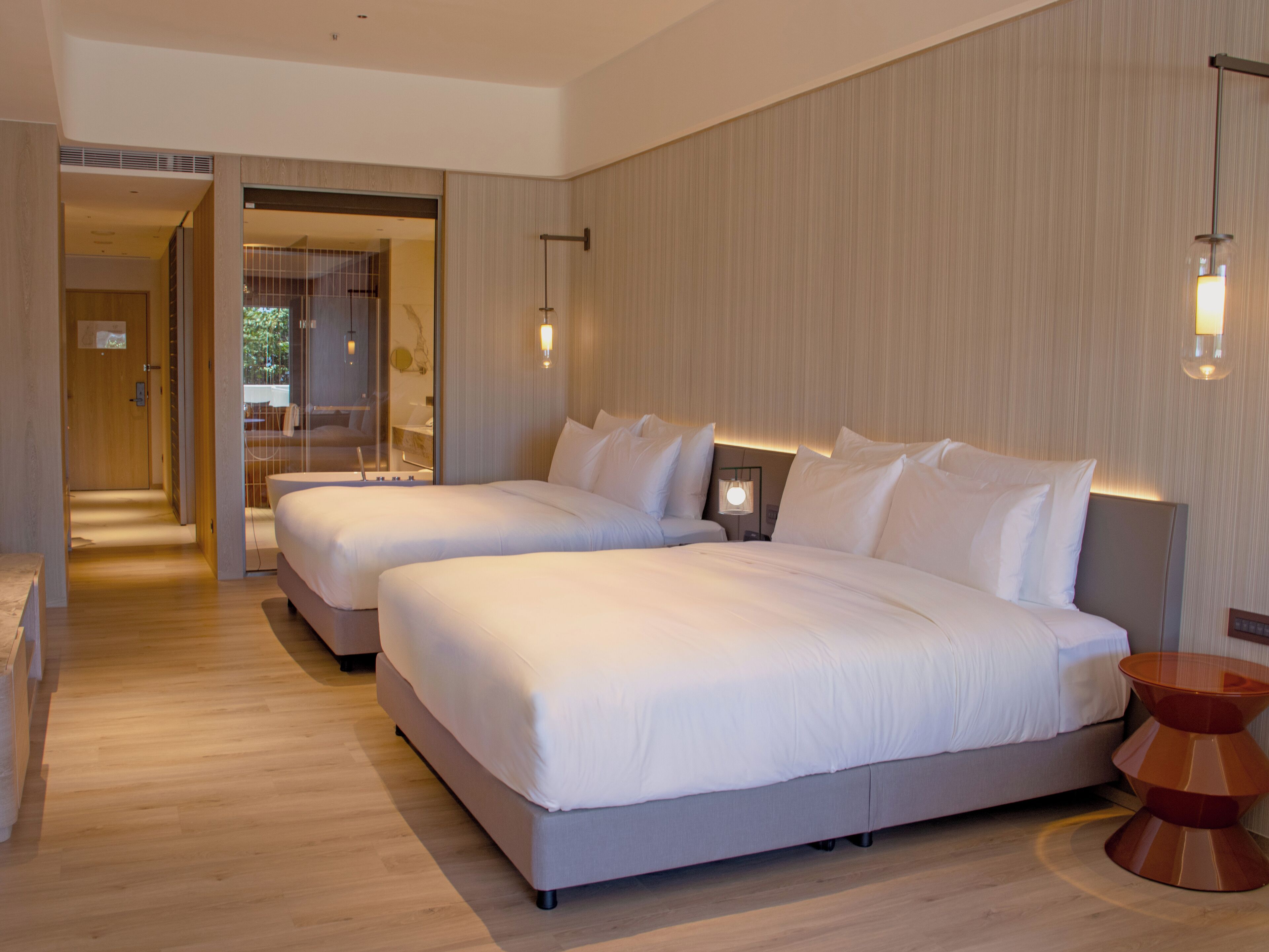 A modern hotel room with two large beds, a wooden floor, and a glass door leading to a bathroom.