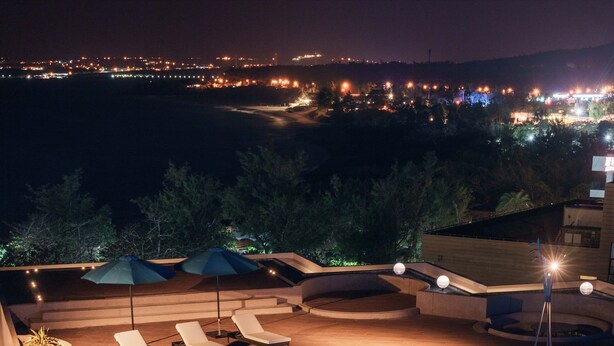 Piscina com bordas iluminadas e aberturas circulares, rodeada por espreguiçadeiras e guarda-sóis, com vista para o horizonte urbano ao fundo.