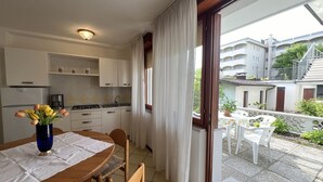 A compact kitchen with white cabinets, a dining table with chairs, and a view of a balcony with outdoor furniture. - Beautiful apartment for 5 guests with A/C, WIFI, TV, patio and pets allowed (Lignano Sabbiadoro)