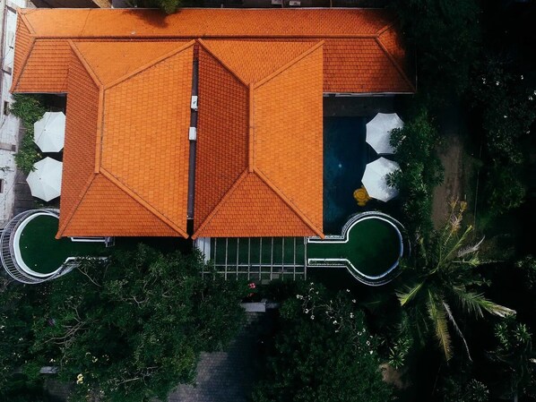 Vista aérea de un edificio con un diseño de techo simétrico, rodeado de exuberante vegetación y una piscina circular.