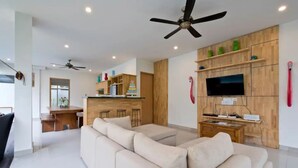 A modern living room with a large beige sofa, a kitchen area with wooden cabinets, a TV mounted on the wall, and ceiling fans. - 6BR Del Mar Villas W/2 Pool near Seminyak Beach (Seminyak)