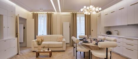 A modern living room with a sofa, dining table, and chairs, a kitchen area with cabinets, and a chandelier.