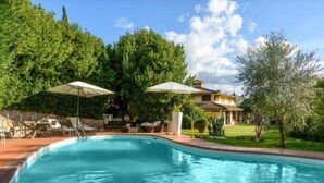 A swimming pool with lounge chairs and umbrellas, surrounded by lush greenery and a house in the background. - Villa Il Guscio by Interhome (Terranuova Bracciolini)