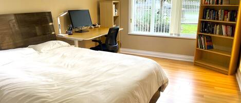 A bedroom with a large bed, a desk with a computer, a bookshelf, and a window with a view of the outdoors.