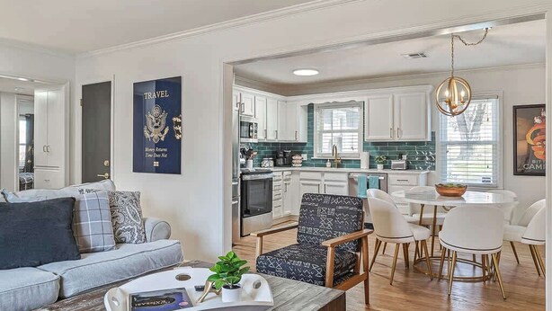 Een moderne woonkamer met een bank, salontafel en een eethoek met stoelen en een tafel. Achtergrond toont een keuken met witte kastjes en een raam.