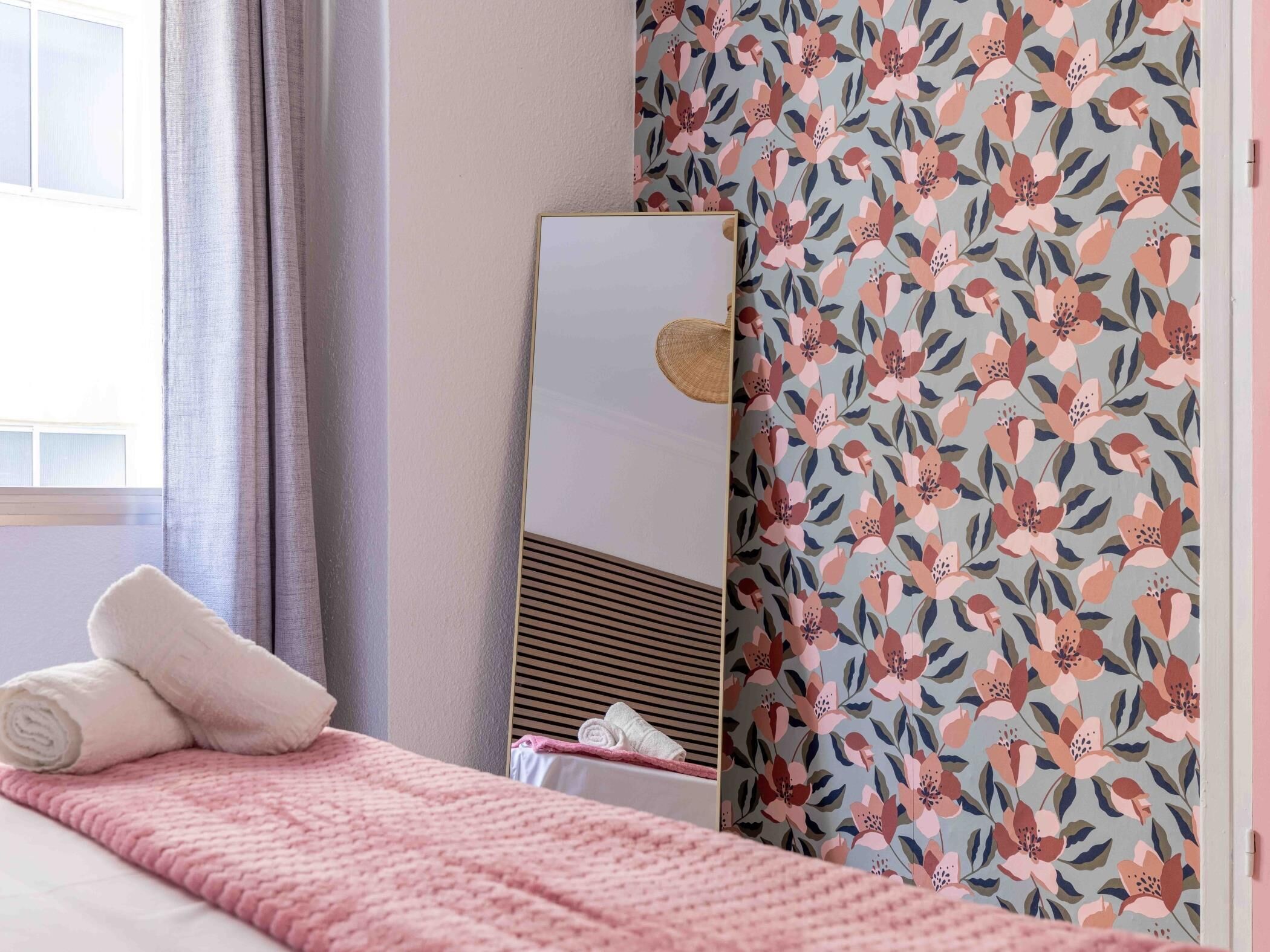 A neatly made bed with a pink blanket and rolled towels, a mirror, and a floral-patterned wall.
