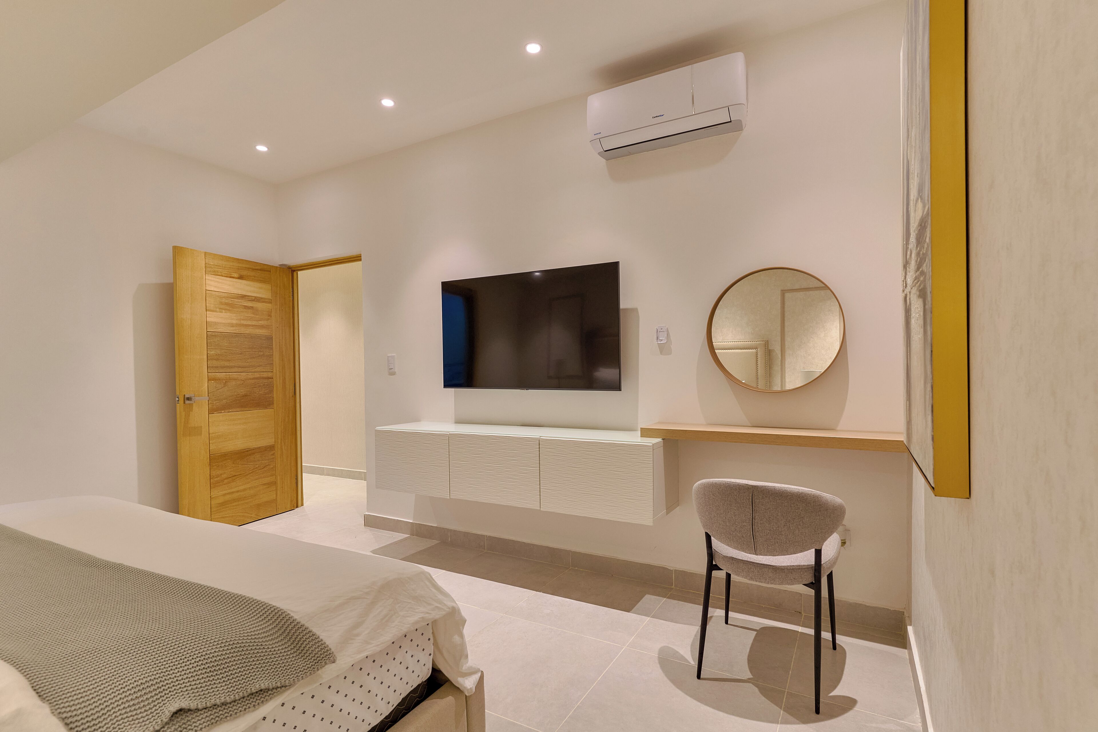 Un dormitorio moderno con una cama, un televisor montado en la pared, un espejo redondo y una silla.