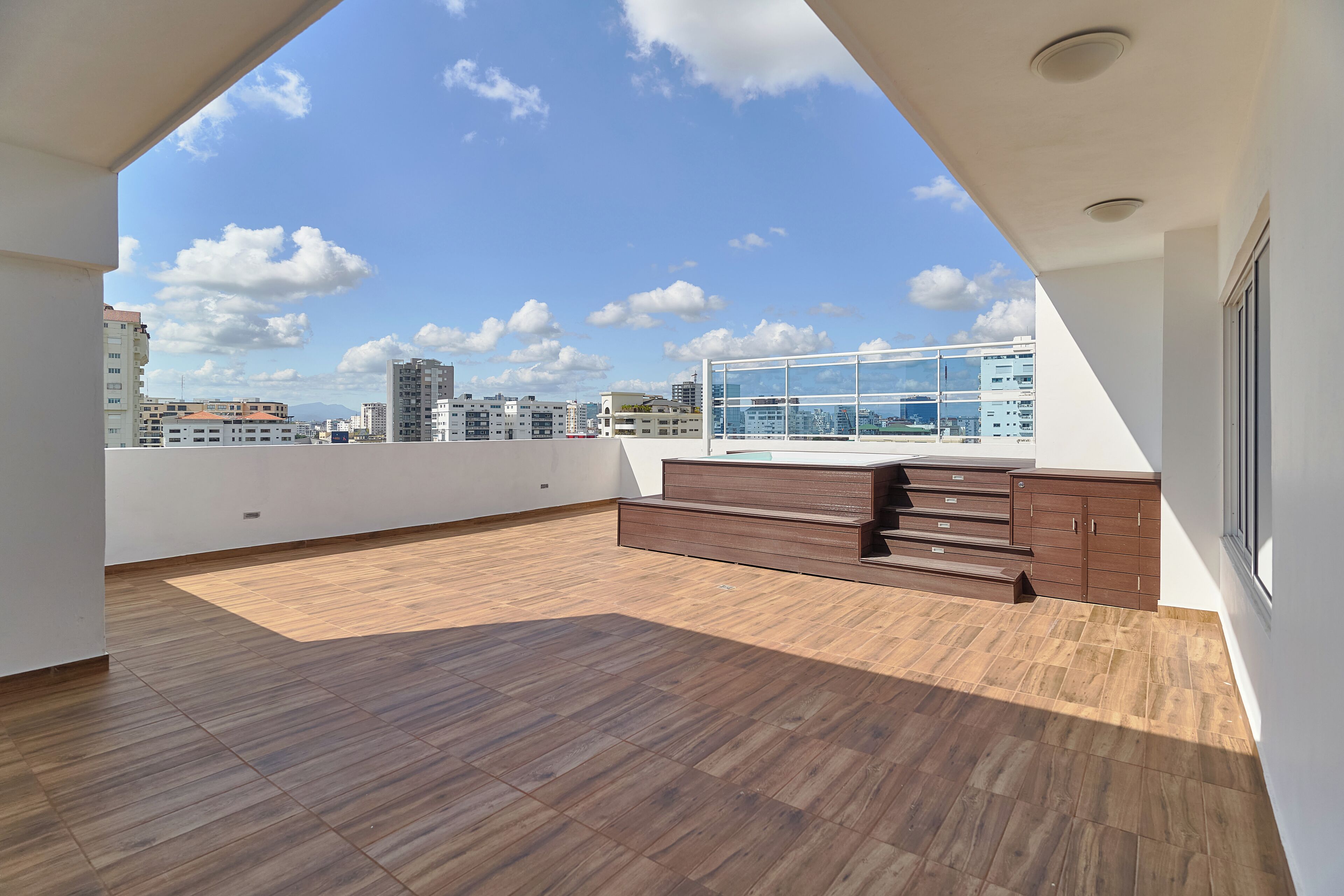 A spacious balcony with a wooden floor, a built-in bench, and a glass railing overlooking a cityscape.