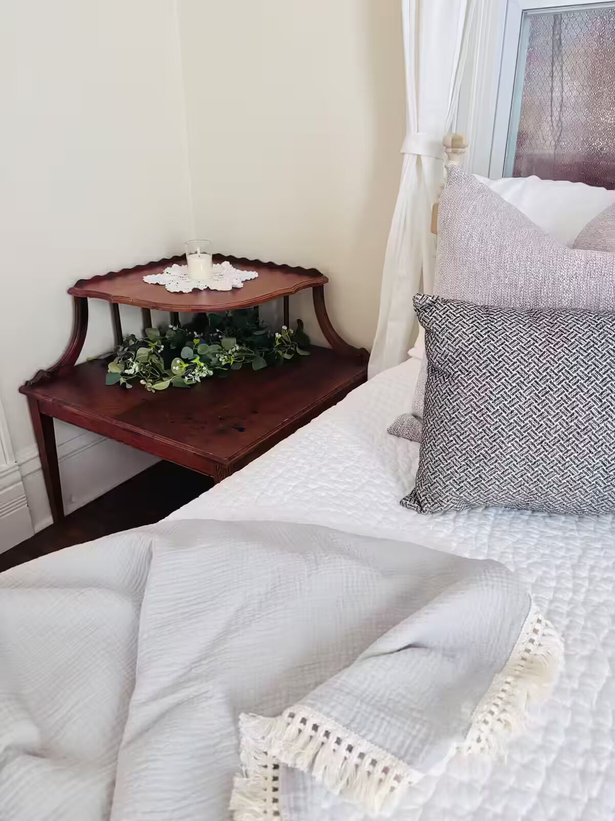A neatly made bed with white bedding, a patterned pillow, and a small wooden nightstand with a candle and greenery.
