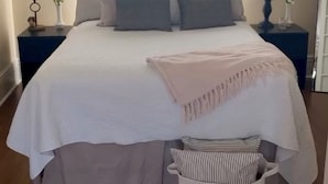 Ein ordentlich bezogenes Bett mit weißer Bettdecke, mehreren Kissen, einer Nachttischlampe und einem Nachttisch mit einer Vase.