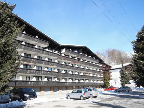 Un hotel de varios pisos con balcones, un estacionamiento y nieve en el suelo.