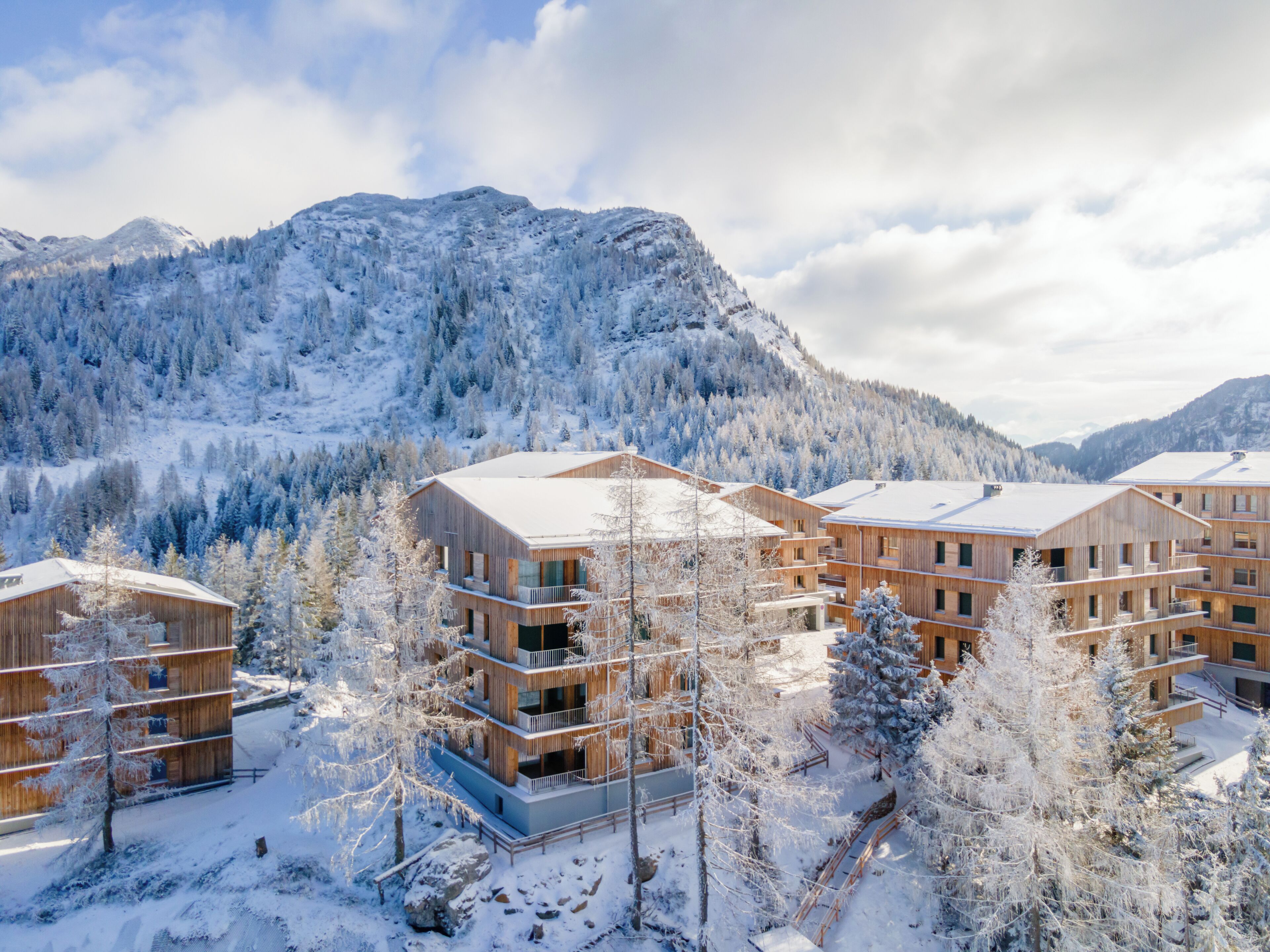 A modern wooden building complex with multiple balconies, surrounded by snow-covered trees and mountains.