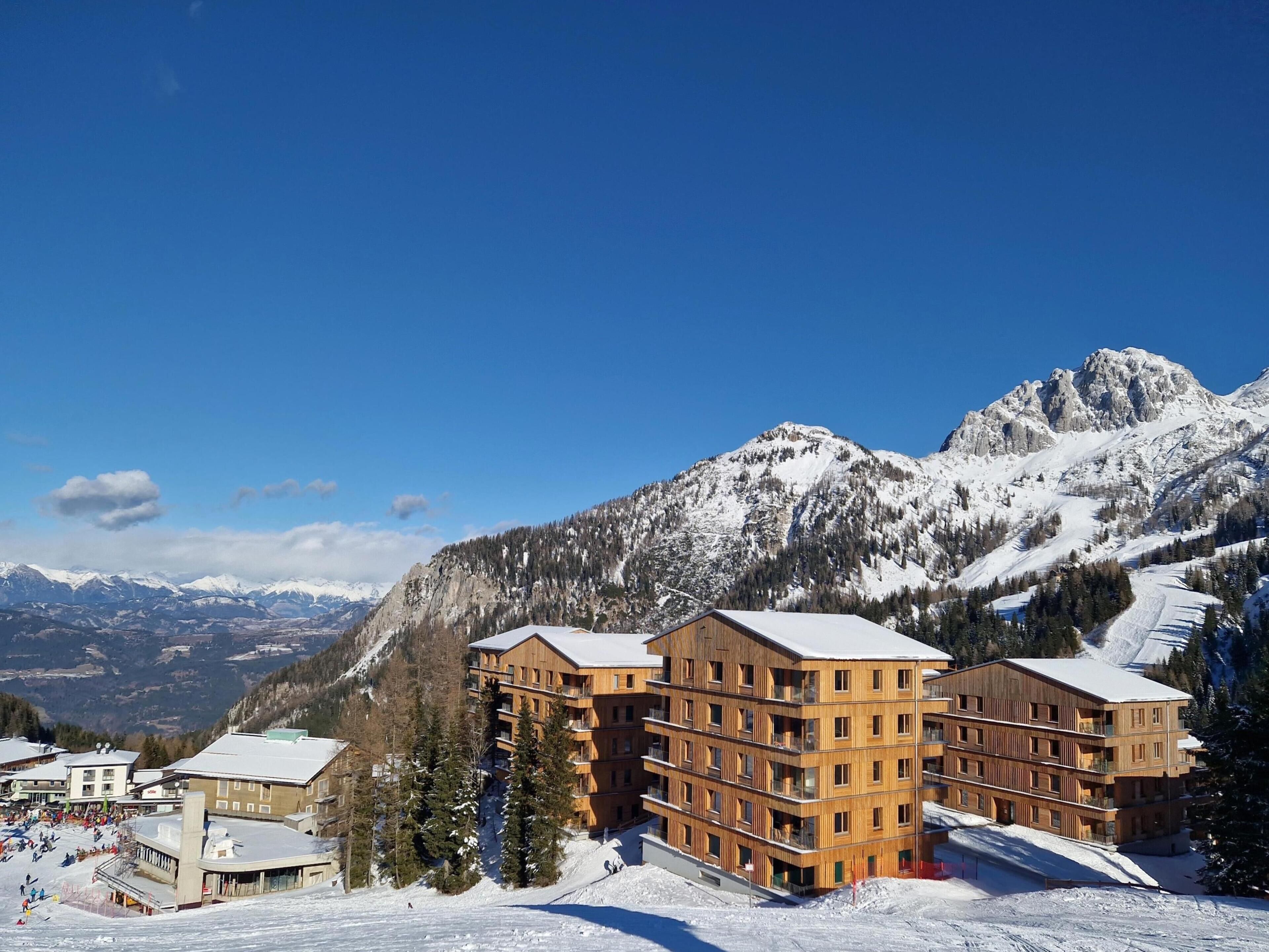 A multi-story wooden building with balconies, surrounded by snow-covered mountains and a clear blue sky.