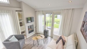 A modern living room with a grey sofa, a white sofa, a coffee table, and a fireplace. There is a television mounted on the wall and a large window with curtains. - Nice house in Zeeland near the Oosterschelde (Kattendijke)