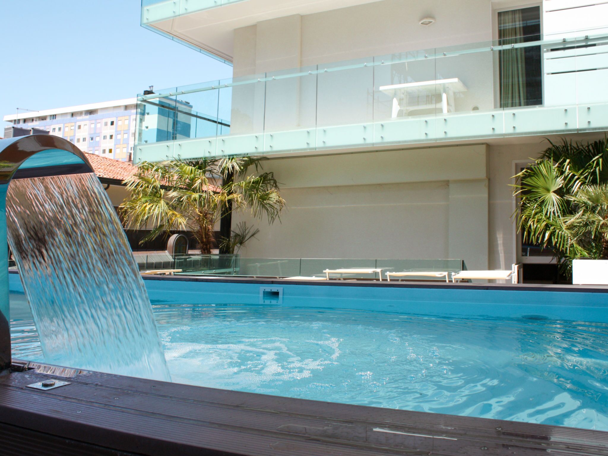 Ein Pool mit Wasserspiel, umgeben von einem modernen Gebäude mit Glasgeländern und Palmen.