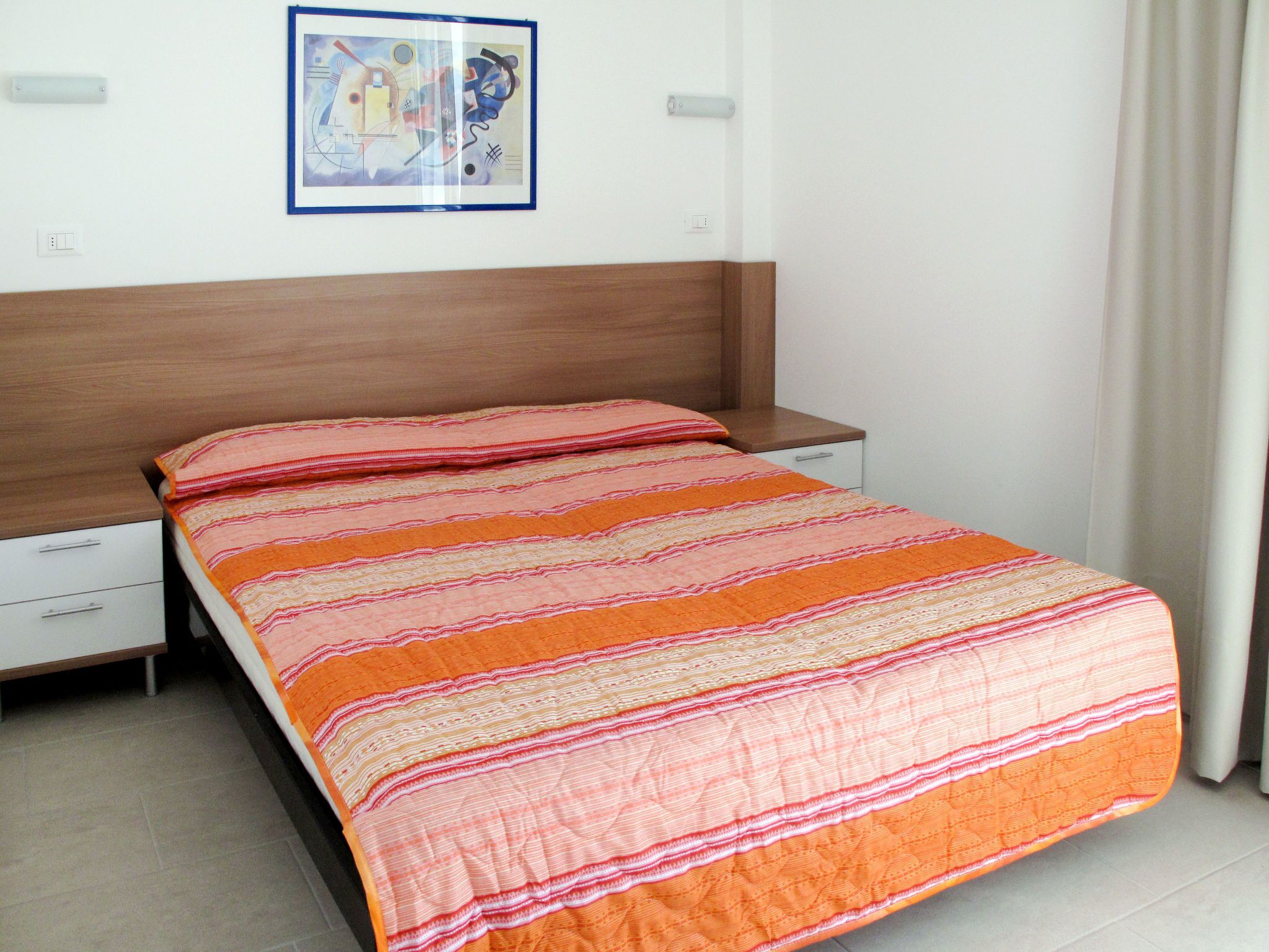 A bedroom with a wooden headboard, a single bed with an orange and white striped bedspread, a nightstand with a drawer, and a framed abstract painting on the wall.