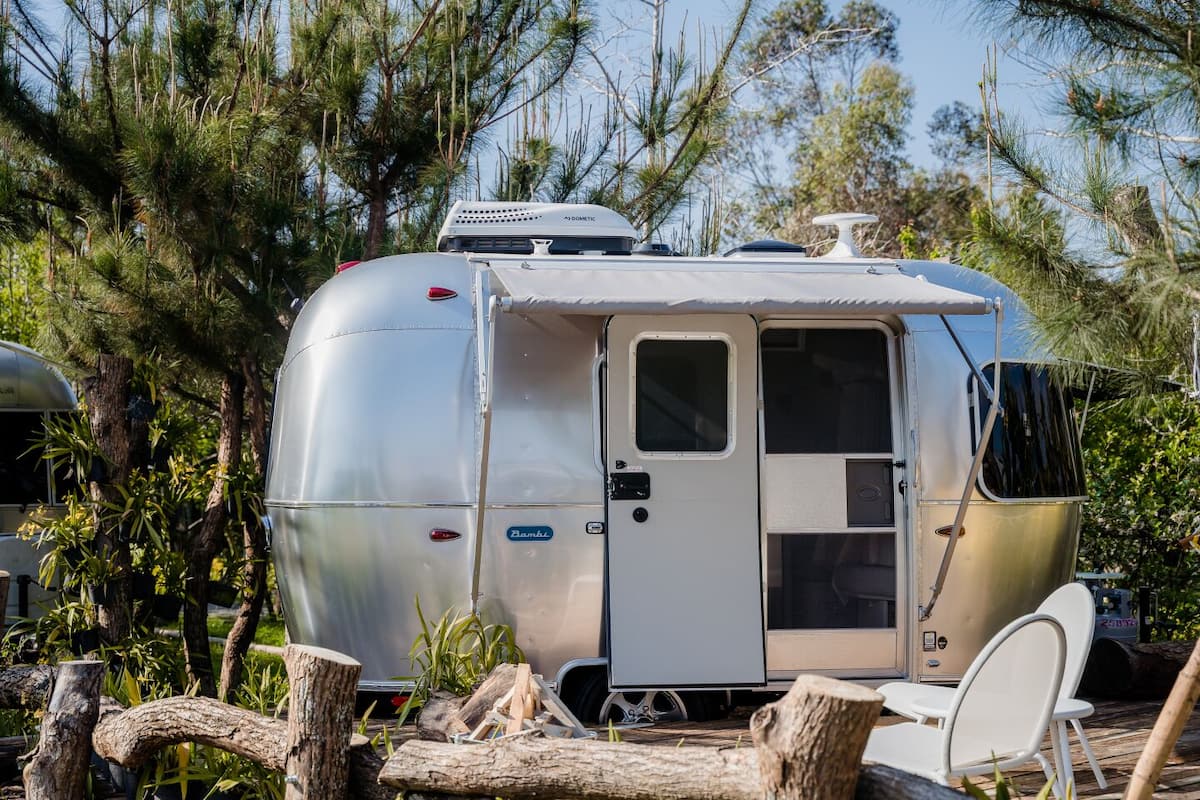 A silver RV with a small attached cabin, surrounded by trees and logs.