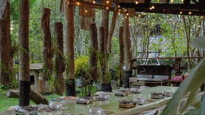 An outdoor dining area with a long table set for a meal, surrounded by trees and plants. - OrangeFarmerlife (Zhuolan)