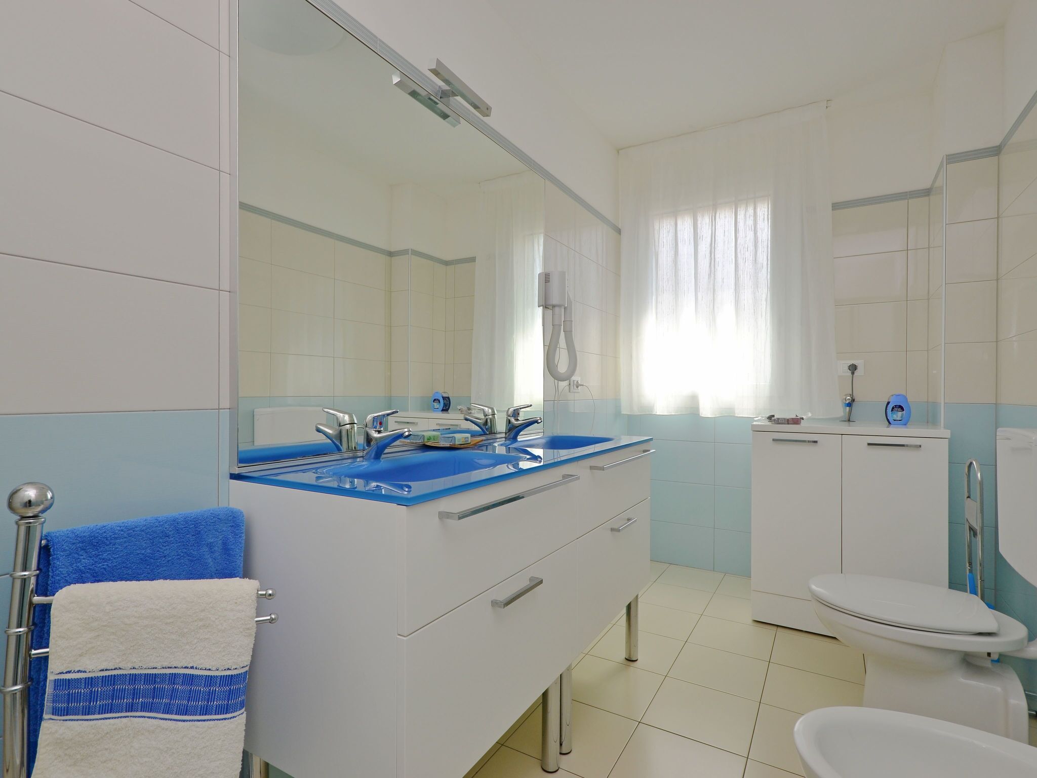 Une salle de bain avec un sol carrelé de blanc et de bleu, un lavabo blanc avec un receveur bleu, un miroir, des toilettes blanches et un porte-serviettes avec une serviette bleue.