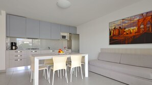 A modern kitchen with grey cabinets, a white dining table with four chairs, a microwave, and a fruit bowl. There is a white sofa and a cityscape painting on the wall.