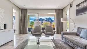 Una sala de estar moderna con una mesa de centro de vidrio, dos sillones, un sofá y vistas a edificios a través de amplios ventanales.