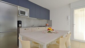 A modern kitchen with a stainless steel refrigerator, a microwave, and a coffee maker. There is a dining table with chairs and a fruit bowl on the table.