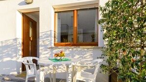 Un área de estar exterior blanca con una mesa, dos sillas, un cuenco de fruta y una planta en maceta.