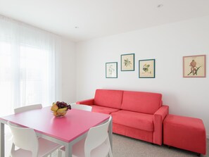 Una habitación luminosa con un sofá rojo, una mesa blanca con un frutero, y tres láminas botánicas enmarcadas en la pared.
